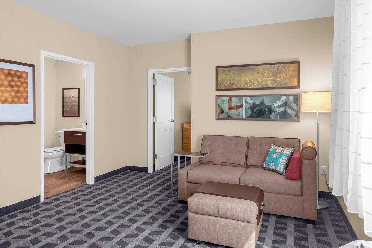 A hotel room with a sofa, ottoman, and a small table. There is a door leading to a bathroom, and a lamp on the right side.