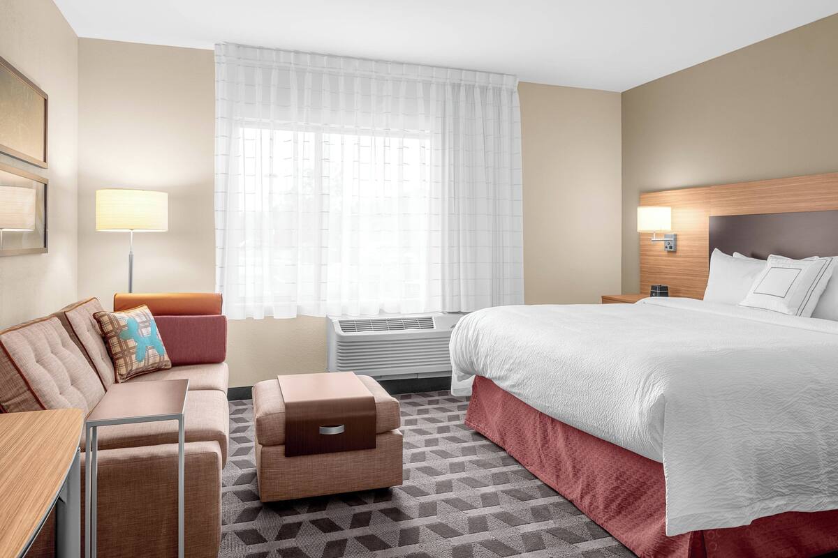 A hotel room with a bed, a sofa, a small table, a lamp, and a window with curtains.
