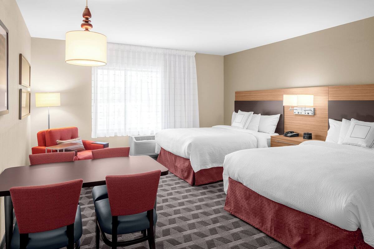 A hotel room with two beds, a desk with chairs, a red sofa, and a large window with curtains.