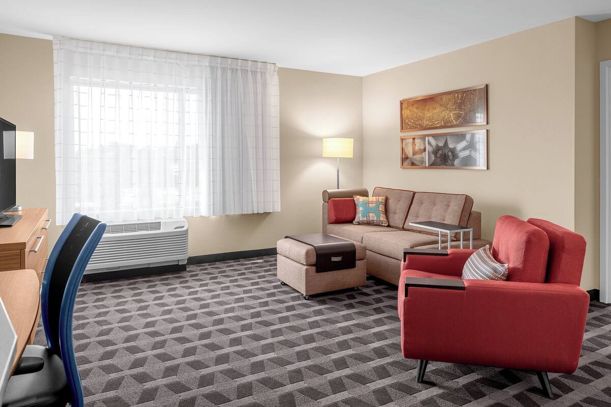 A hotel room with a sofa, armchair, and ottoman, a desk with a chair, a lamp, and a window with curtains.