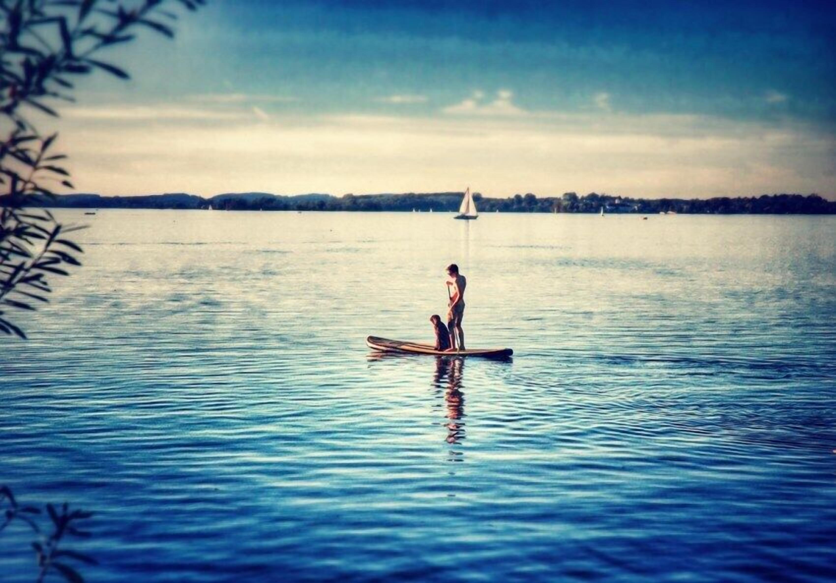 A calm lake with a sailboat in the distance and a person standing on a paddleboard.