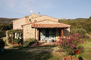 Una casa de piedra con techo de tejas, un porche cubierto con mesa y sillas, y un jardín con plantas y flores en macetas.