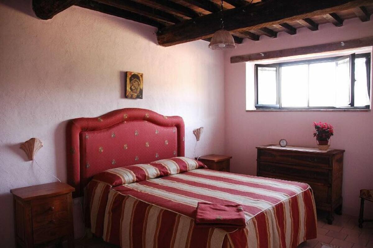 Un dormitorio con una cama tapizada en rojo, un aparador de madera, una mesita de noche, una lámpara de pared y una ventana con vistas al exterior.