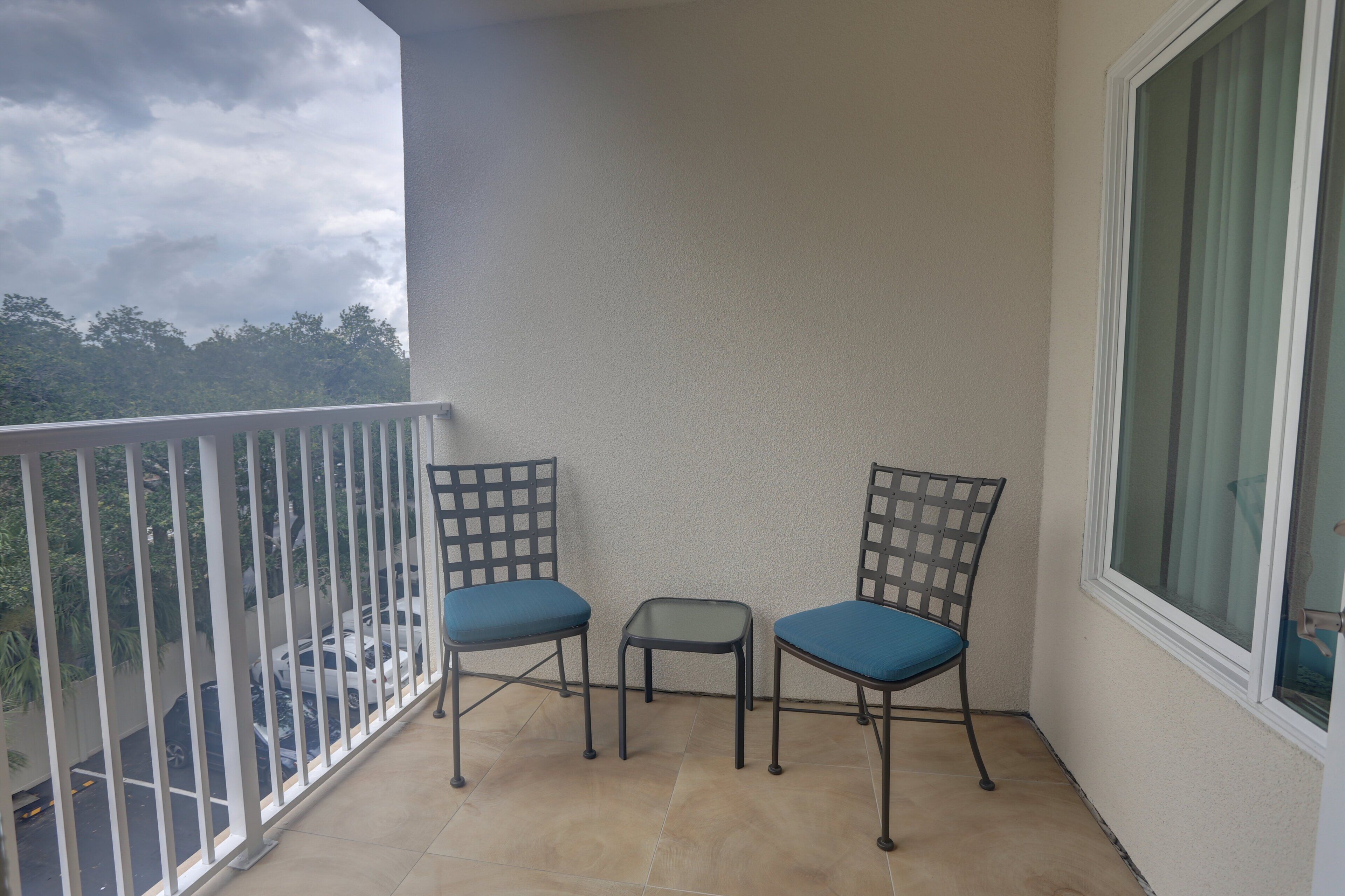 Ein Balkon mit zwei Stühlen und einem kleinen Tisch, einem weißen Geländer und Blick auf parkende Autos und Bäume.