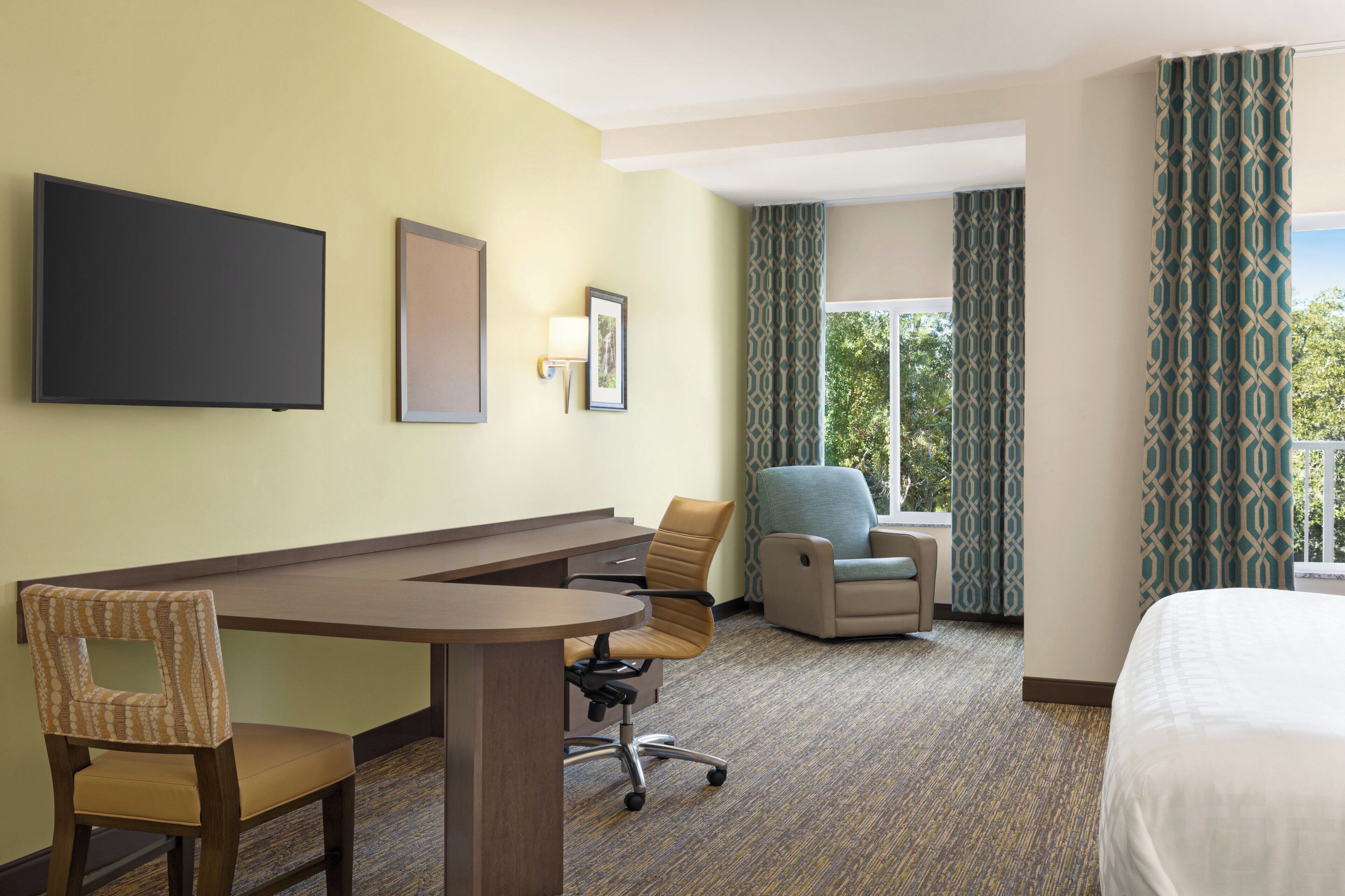 A hotel room with a bed, a desk with a chair, a sofa, a TV, and a window with curtains.
