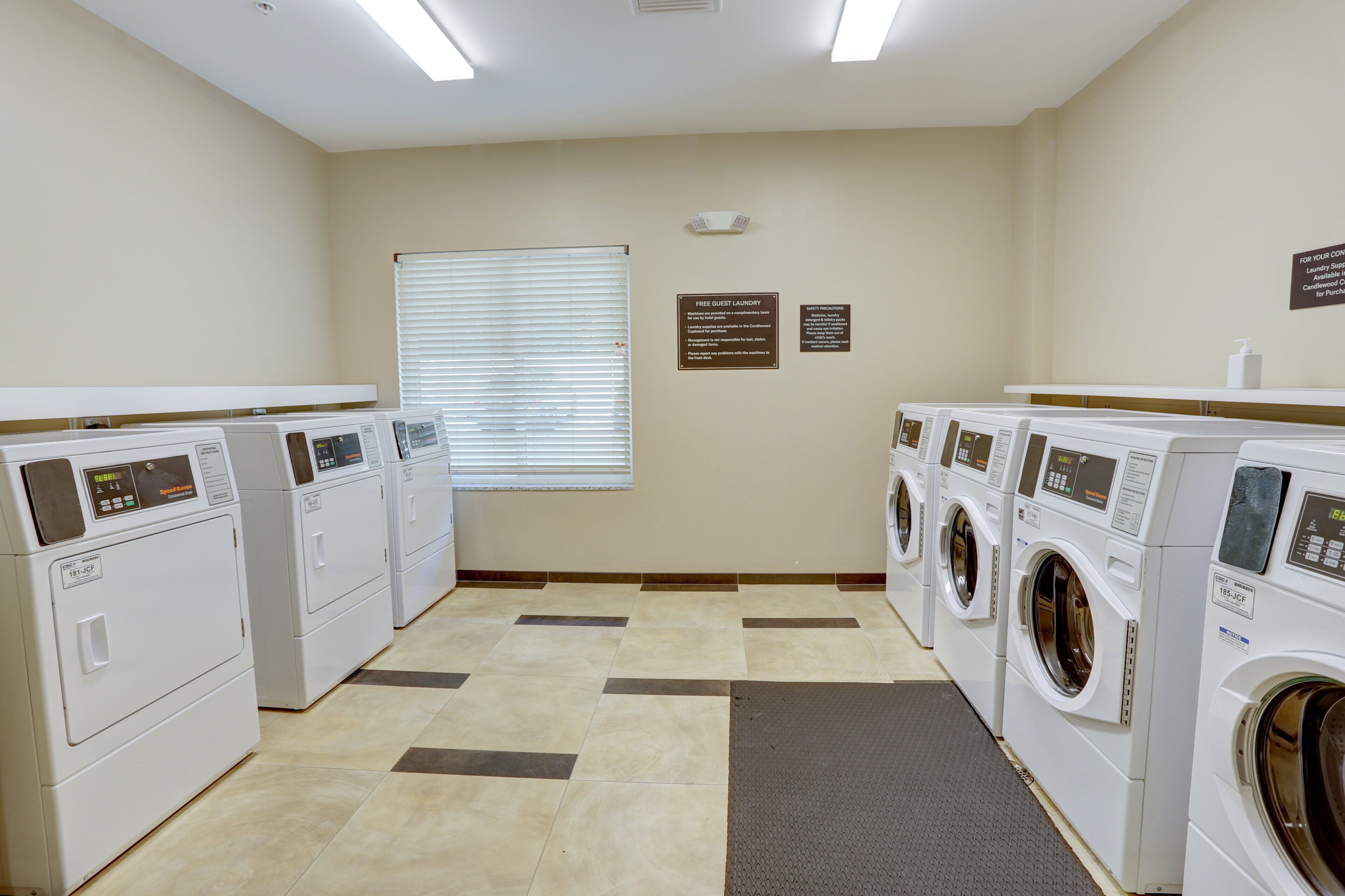 Une buanderie équipée de plusieurs machines à laver et de plusieurs sèche-linge, une fenêtre avec des stores et des panneaux d’information fixés aux murs.