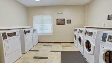A laundry facility with multiple washing machines and dryers, a window with blinds, and informational signs on the walls.