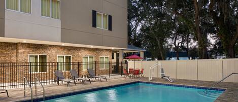 A swimming pool with lounge chairs and a covered seating area, surrounded by a brick building and a fence.