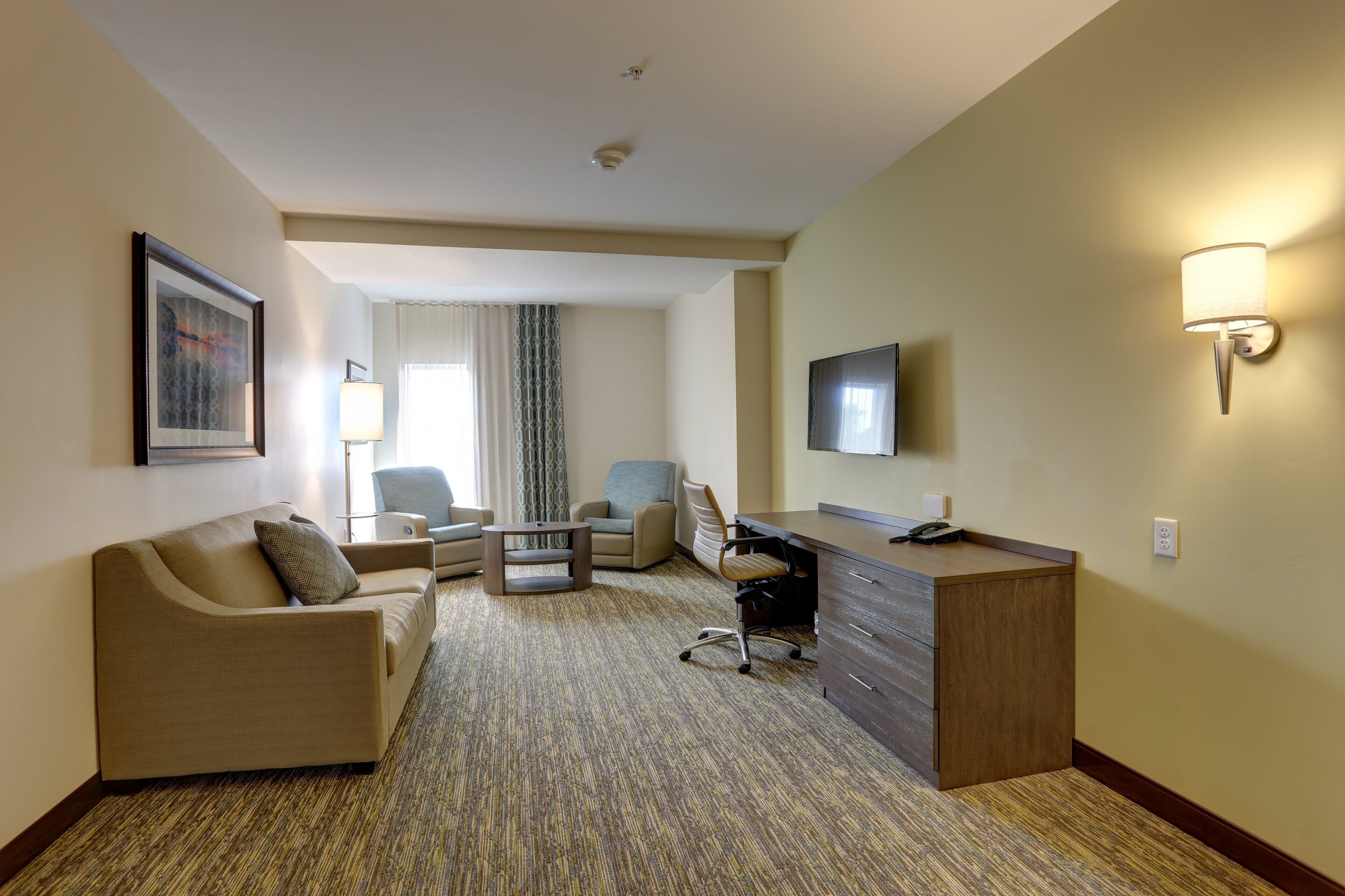 A hotel room with a sofa, two armchairs, a desk, a television, and a patterned carpet.