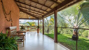 A covered patio with a wooden bench, potted plants, and a view of a lush garden.