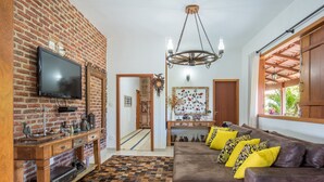 Un salon avec un mur en briques, un téléviseur, une console en bois, un tapis à motifs, un canapé marron avec des coussins jaunes et un lustre.
