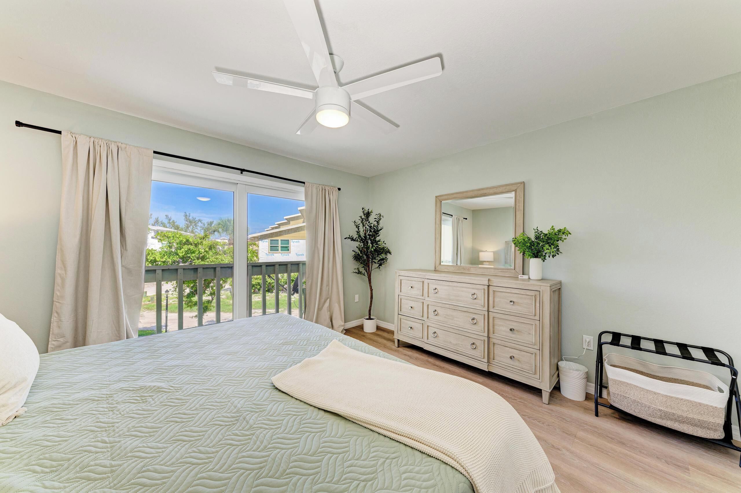 A bedroom with a bed, a dresser, a mirror, a ceiling fan, and a balcony with curtains.