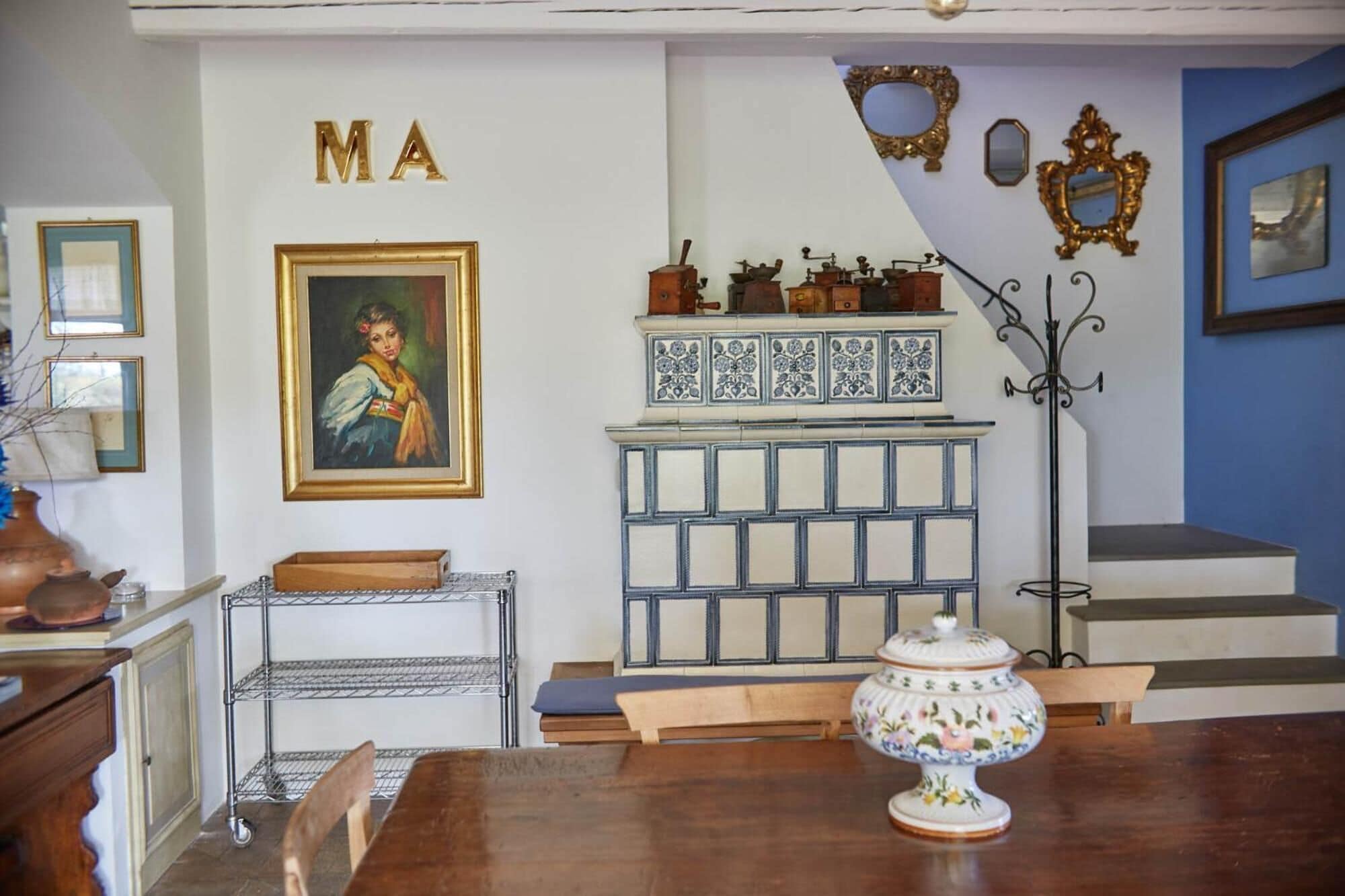 Un'area pranzo con un tavolo di legno, un mobiletto a mensole metalliche, una decorativa anfora e un pensile con piastrelle blu e bianche.