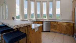 Une cuisine dotée d’un grand îlot central, de meubles en bois, d’un lave-vaisselle en acier inoxydable et de nombreuses fenêtres offrant une vue sur les montagnes.