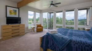 Une chambre avec un grand lit, un téléviseur posé sur une commode en bois, un ventilateur de plafond et une vue sur un paysage verdoyant grâce à de grandes fenêtres.