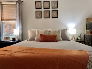Quarto com cama, almofadas, mesa de cabeceira, abajur, janela com persianas e quadros nas paredes.