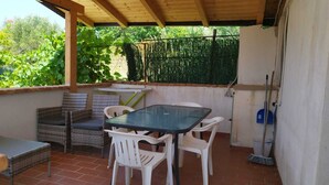 Une terrasse couverte avec une table et des chaises, une petite table avec une chaise, et un balai appuyé contre le mur.