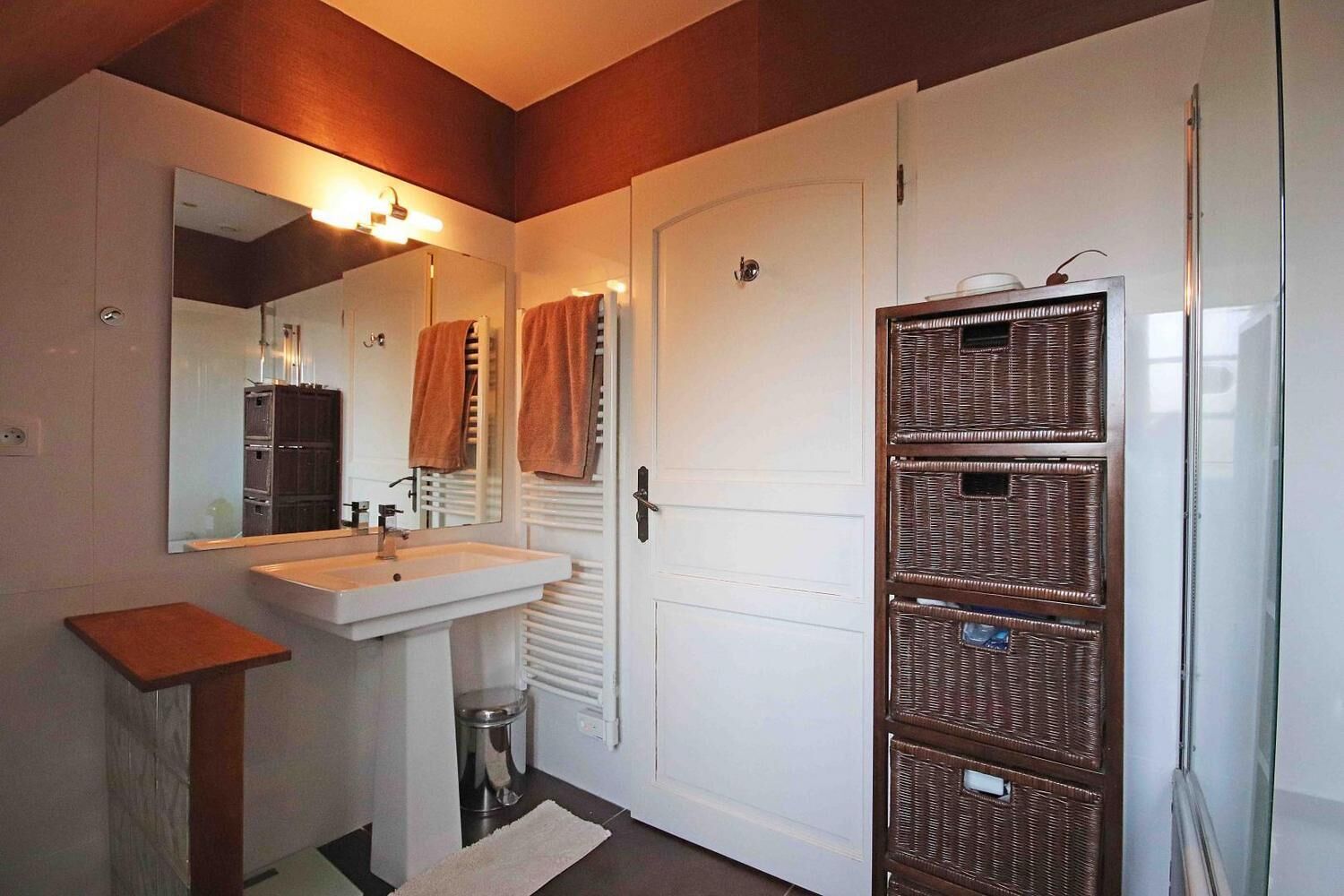 A bathroom with a white sink, mirror, and towel rack. There is a door with a white handle and a wicker basket storage unit.