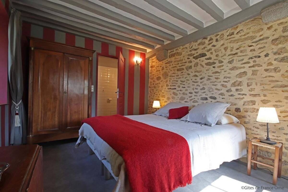 Ein Schlafzimmer mit Steinwand, Holzschrank, Bett mit roter Decke und einer Nachttischlampe.