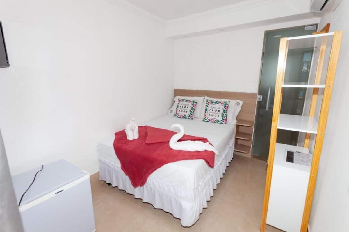 Quarto bem arrumado com cama, travesseiros, manta vermelha e uma pequena mesa branca.
