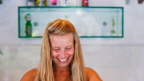 A person in a red top cutting food on a wooden table with a knife and a bowl of greens, in a kitchen with a glass shelf in the background. - Macaco Surf Hostel (Tibau do Sul)