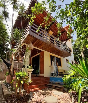 A two-story house with a balcony, surrounded by tropical plants and a stone pathway. - Macaco Surf Hostel (Tibau do Sul)