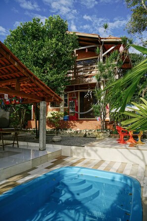 A pool area with a wooden pergola, a brick building with a balcony, and tropical plants. - Macaco Surf Hostel (Tibau do Sul)
