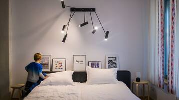 A bedroom with a bed, a hanging light fixture, framed pictures on the wall, and a window with curtains.
