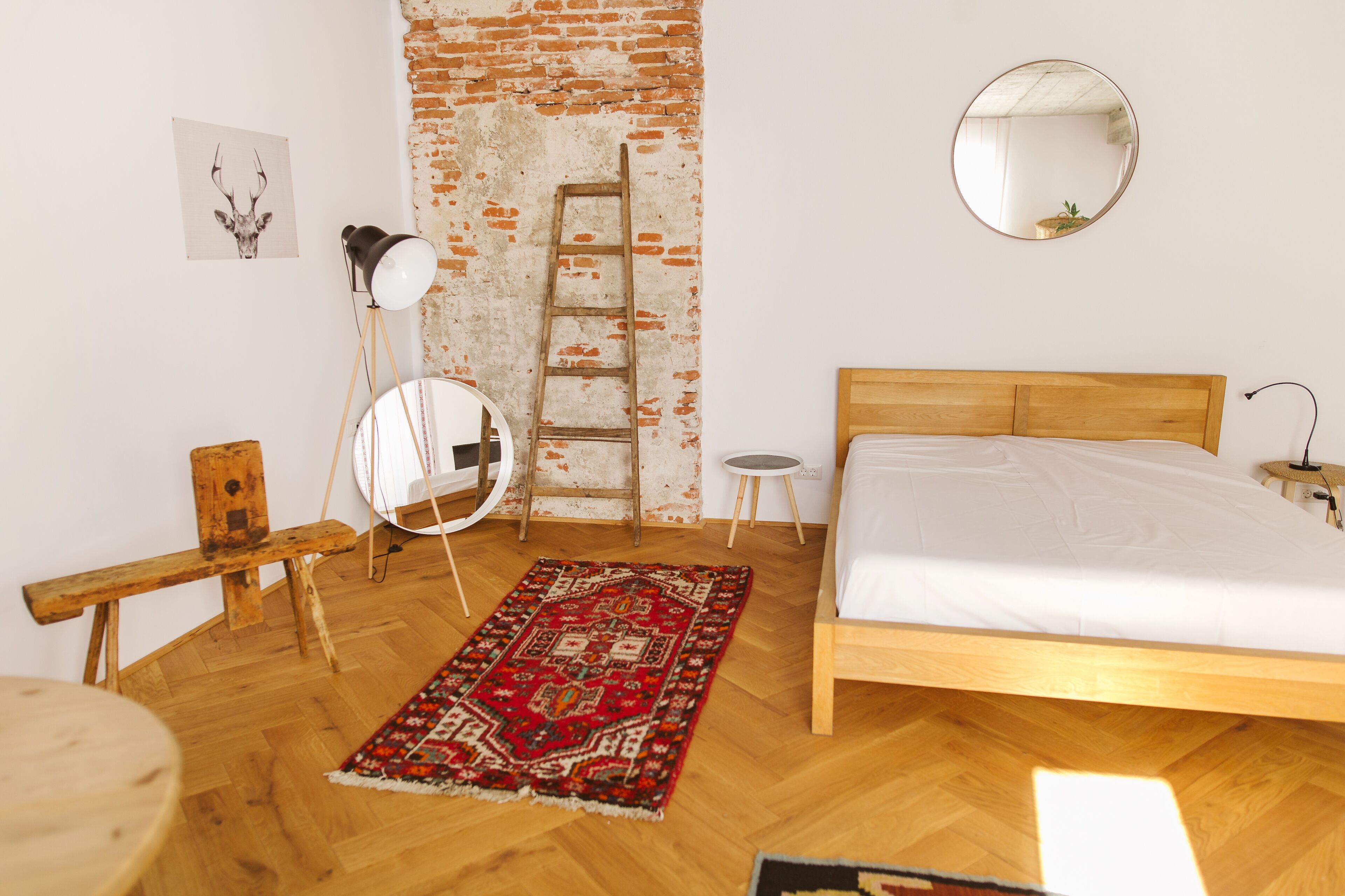 Un dormitorio con una cama de madera, un tapete rojo estampado, un banco de madera, un espejo redondo, una escalera y una mesita.