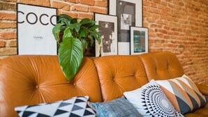 A leather sofa with a large green plant and patterned cushions against a brick wall with framed pictures.