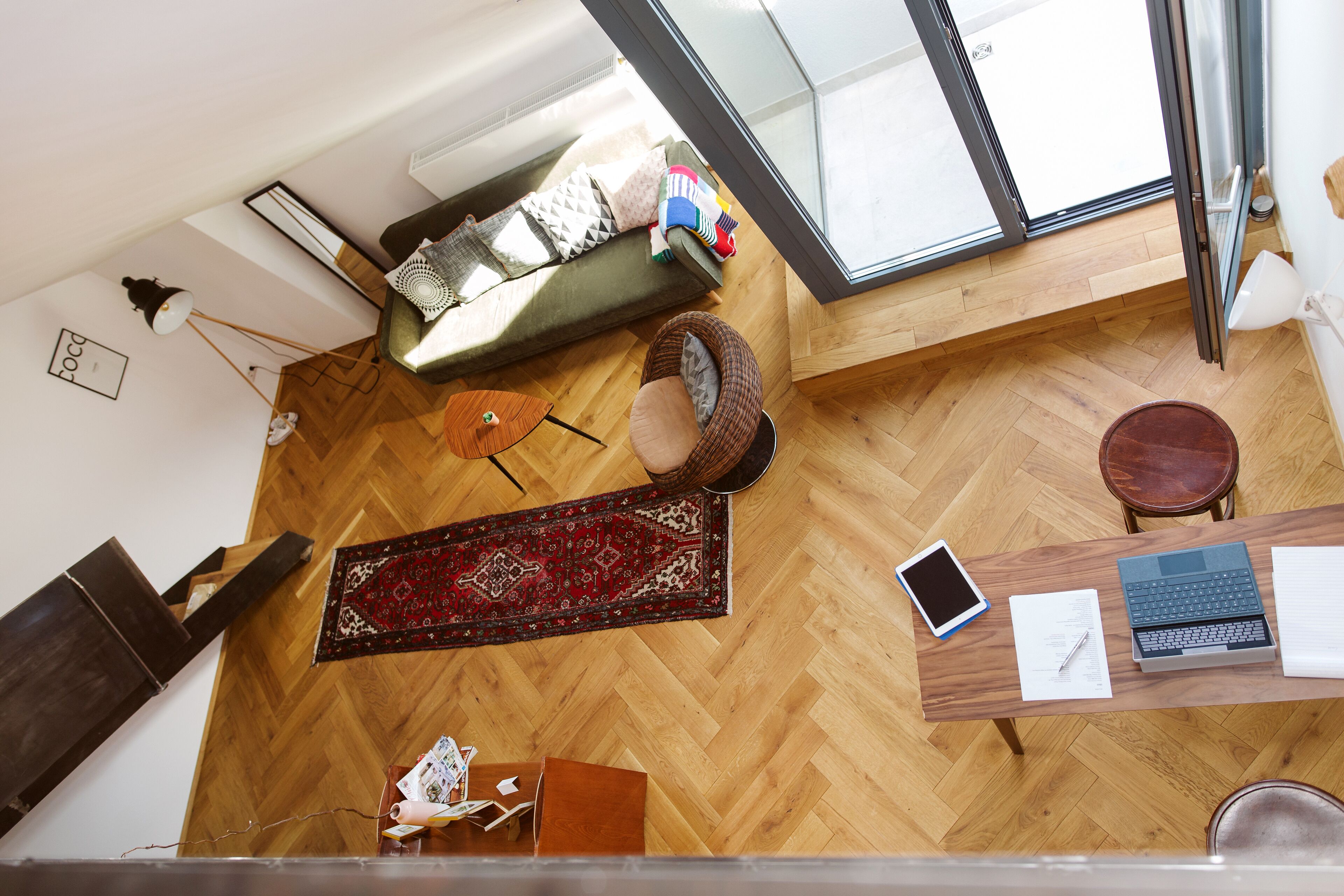 A room with a wooden floor, a sofa, a small rug, a desk with a laptop, a tablet, and a chair.