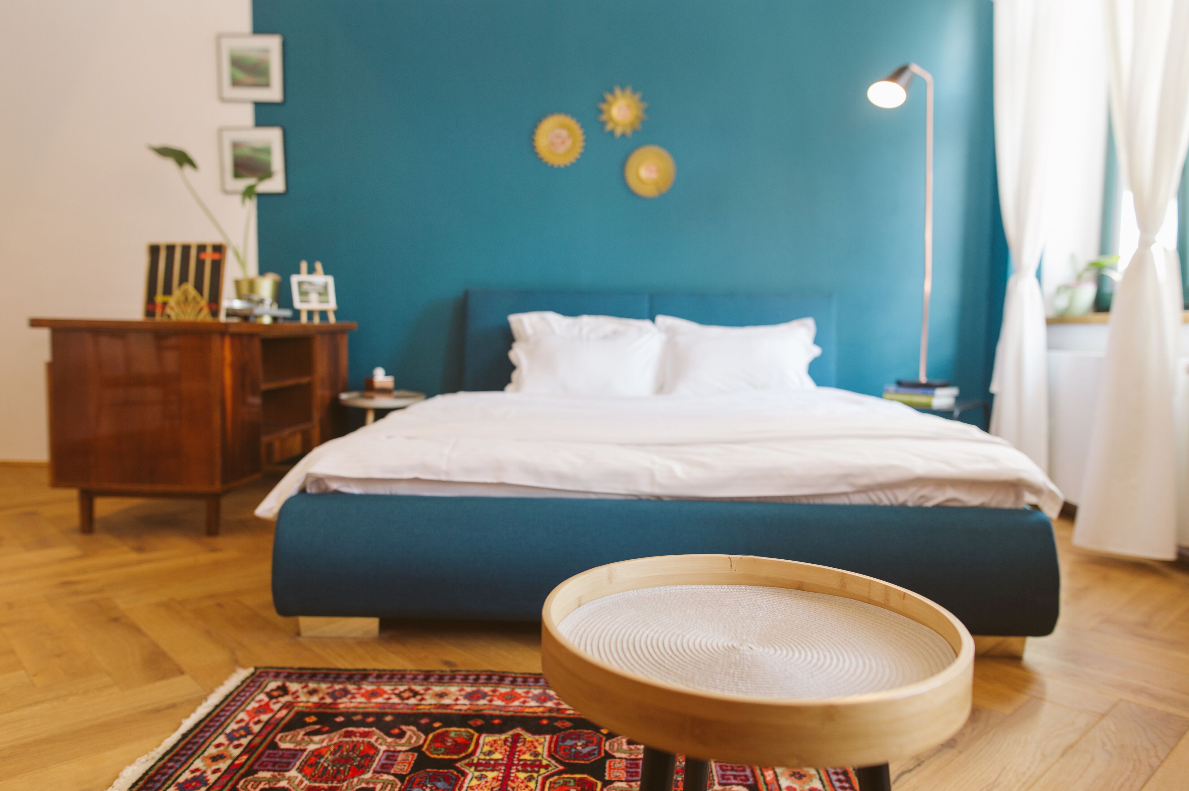 A bedroom with a blue bed, white pillows, a wooden nightstand, a lamp, and a decorative wall piece.