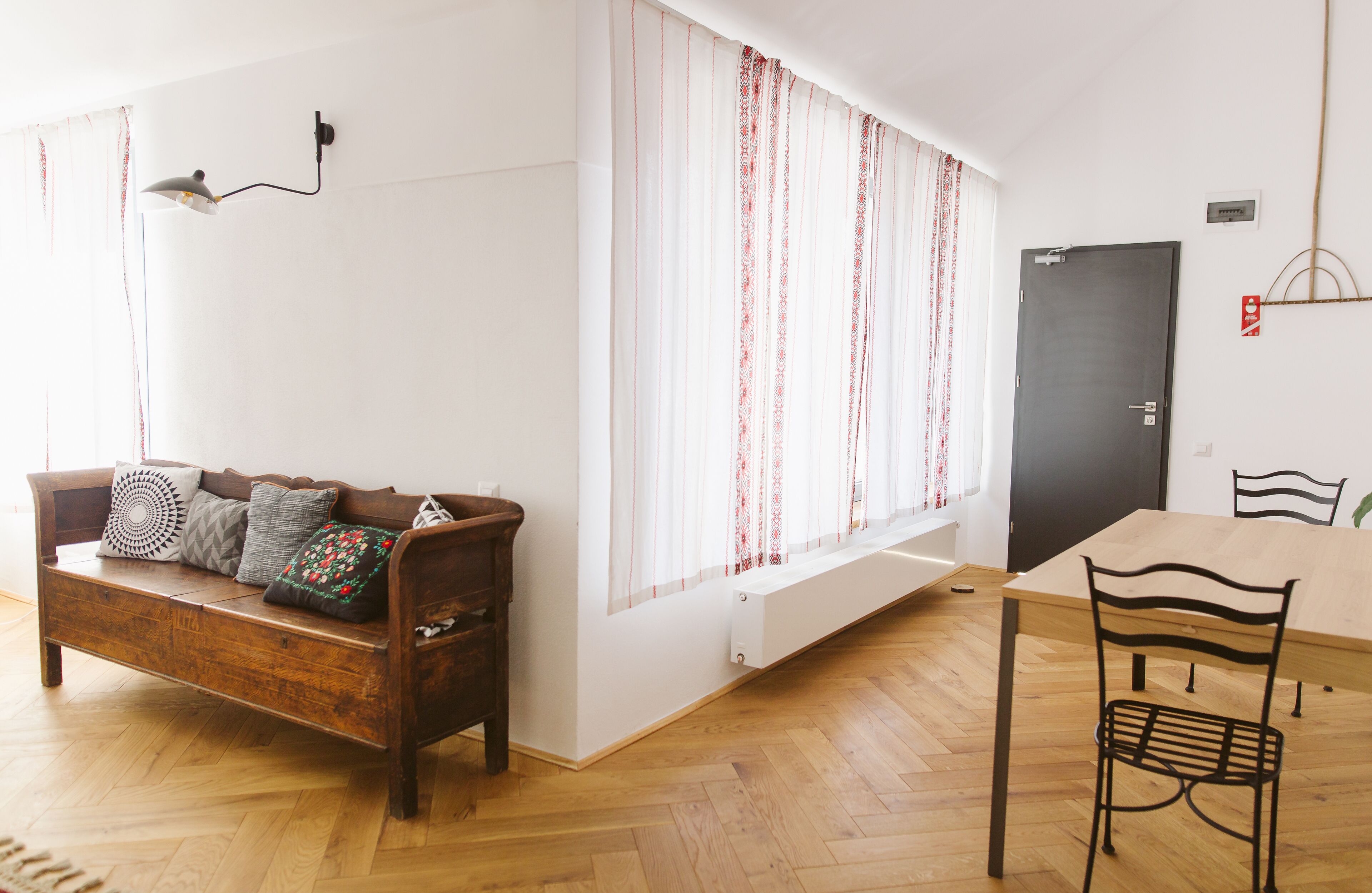 A room with a wooden floor, a wooden sofa with patterned cushions, a dining table with chairs, and a window with curtains.