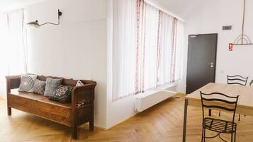 A room with a wooden floor, a wooden sofa with patterned cushions, a dining table with chairs, and a window with curtains.