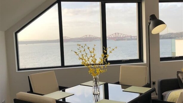 Uma sala de jantar com uma mesa e cadeiras, um vaso com flores amarelas e vista para uma ponte sobre a água através de grandes janelas.