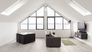 Amplo quarto no estilo loft, com grandes janelas no teto, uma cama, uma escrivaninha e uma televisão.