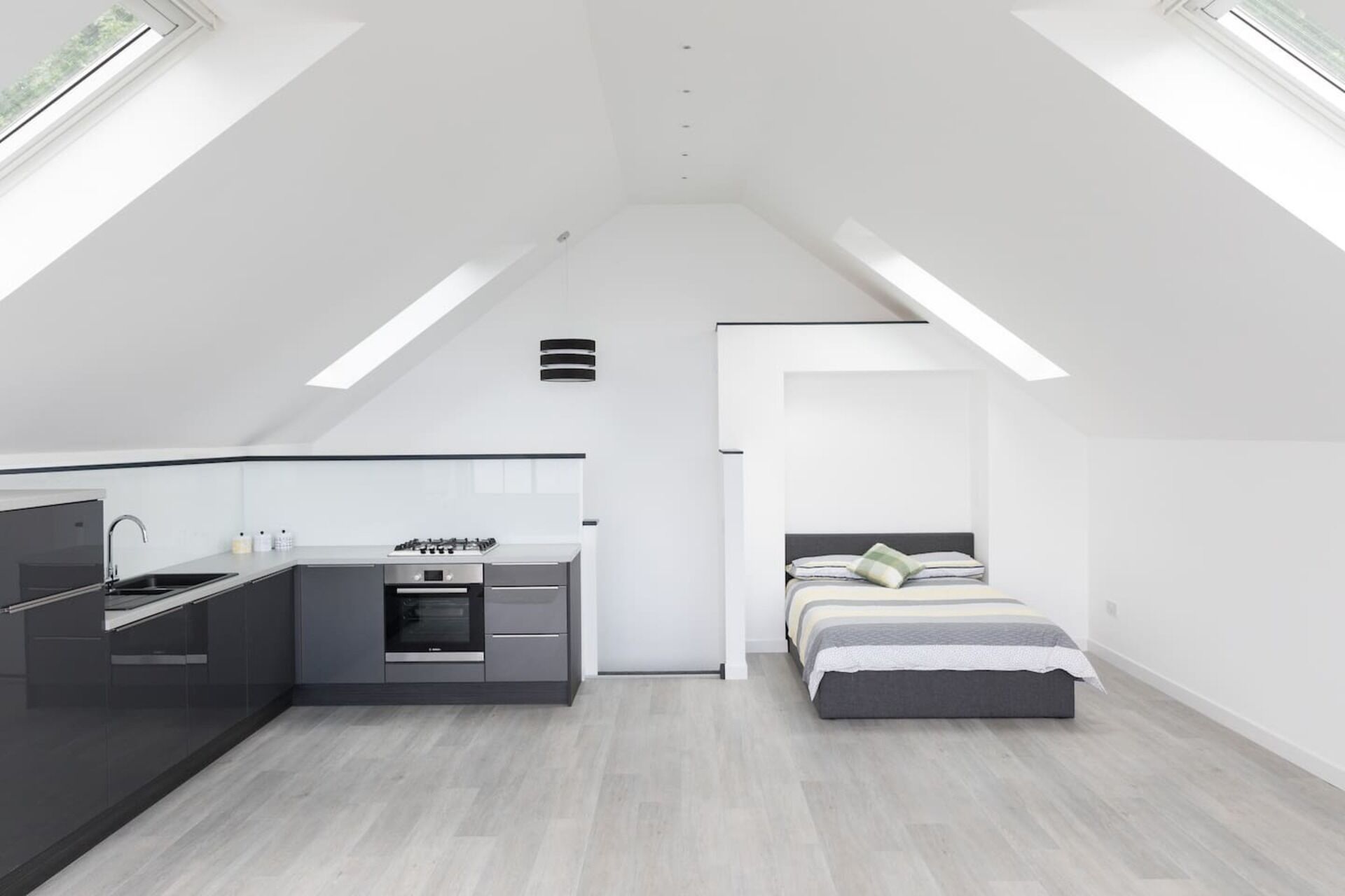 A modern loft bedroom with a built-in kitchen, a bed with a gray headboard, and skylights.