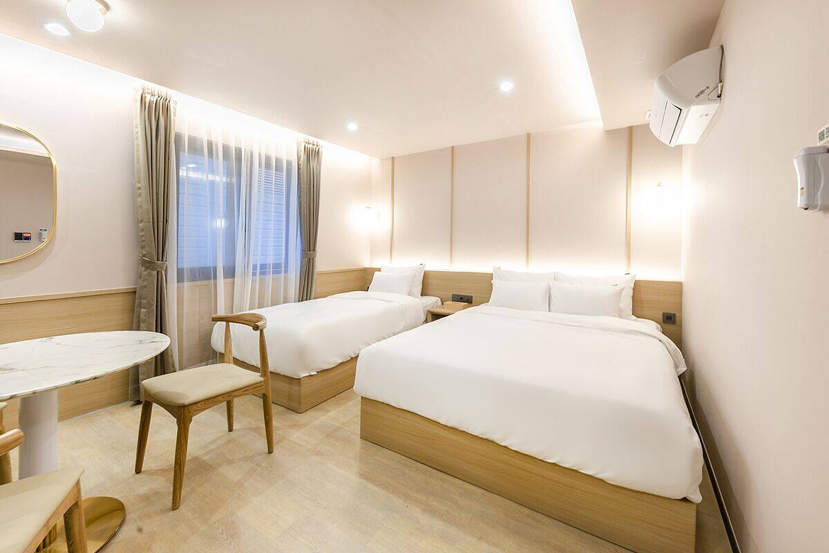 A hotel room with two beds, a small table, a chair, a mirror, and a window with curtains.