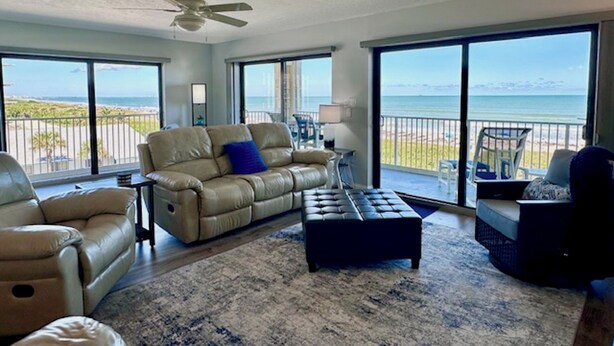 Un salon spacieux doté d’une grande fenêtre donnant sur la plage, d’un ventilateur de plafond et d’un canapé en cuir agrémenté d’un coussin bleu.