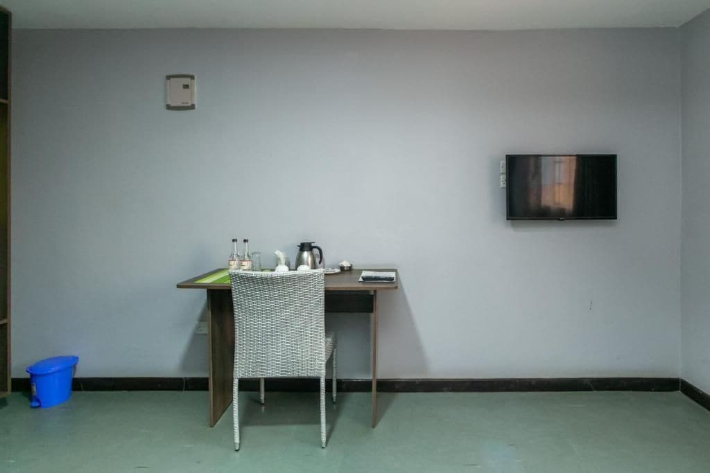 A room with a single bed, a small table with a chair, a television mounted on the wall, and a blue bucket on the floor.