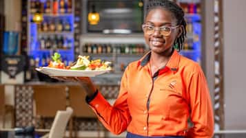 Una persona con uniforme naranja sostiene un plato con comida, de pie en una zona de bar con estantes llenos de botellas y una barra.