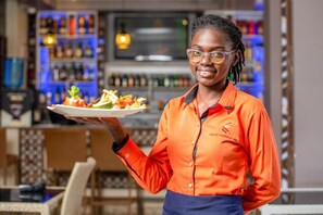 A person in an orange uniform holding a plate with food, standing in a bar area with shelves of bottles and a bar counter.