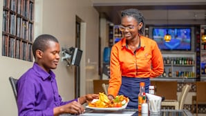 A person serving food to another person at a table in a restaurant setting. - Kings Premier Inn (Mlolongo)