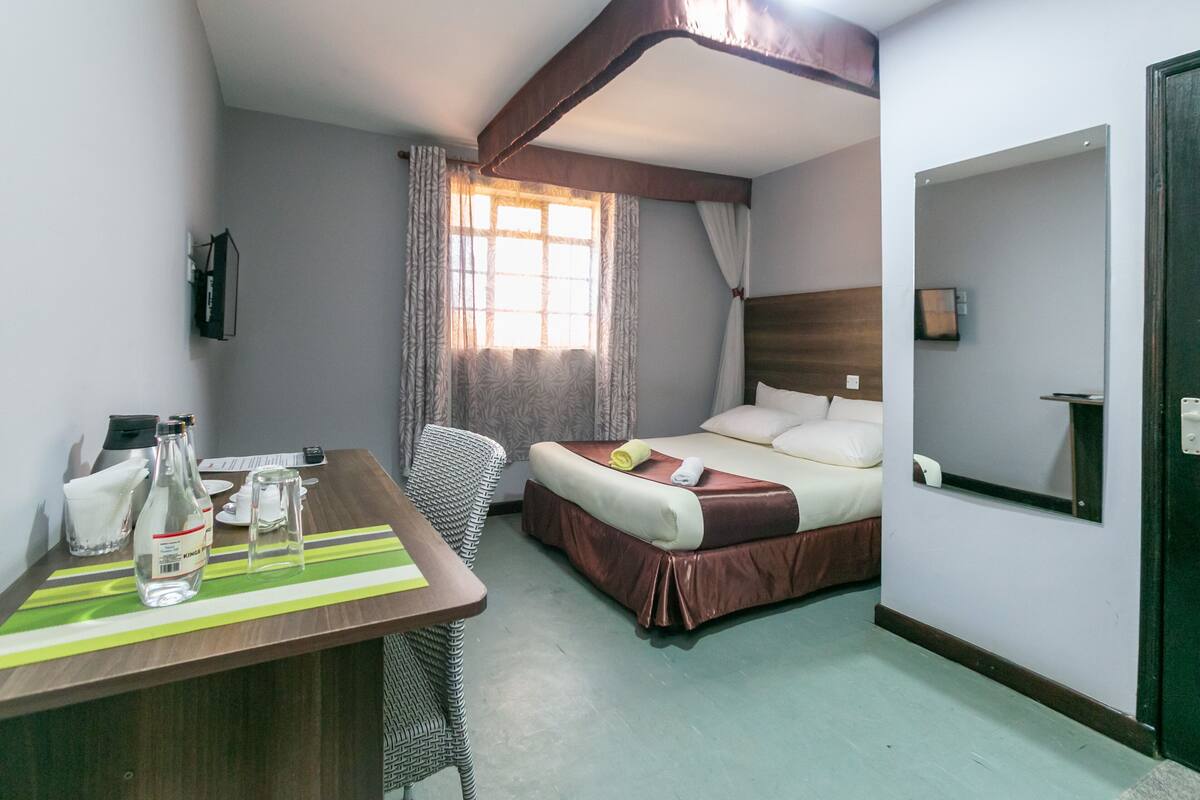 A hotel room with a bed, a desk with a chair, a window with curtains, and a mirror on the wall.