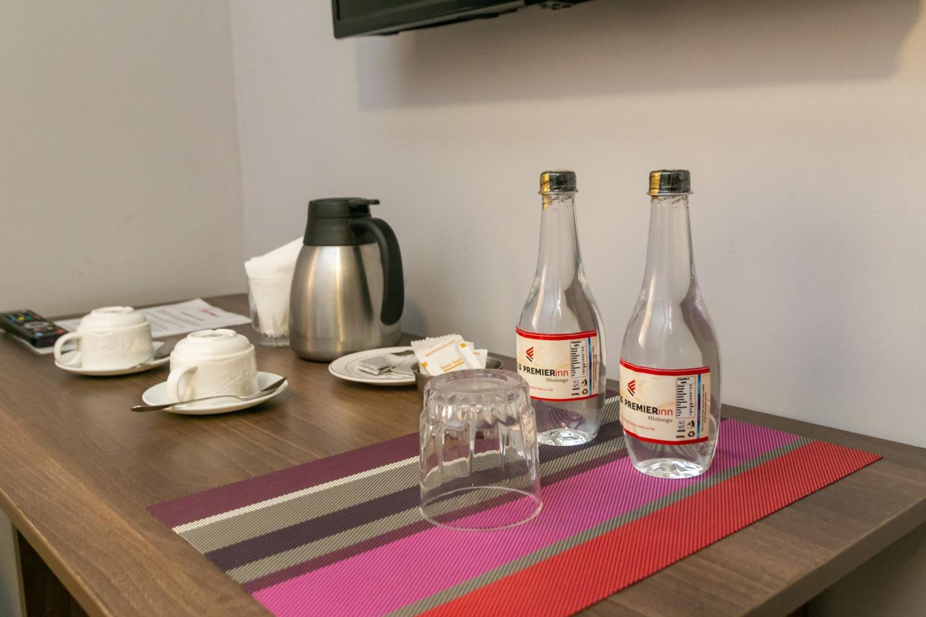 A hotel room with a wooden table, two bottles of water, a glass, a kettle, and a striped placemat.