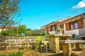 A stone house with a tiled roof, wooden shutters, and a garden area with plants and a stone bench.