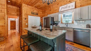 L’intérieur d’un chalet en bois comprend un îlot de cuisine, des tabourets de bar, un lustre et un panneau sur lequel on peut lire le mot « rassemblement ».