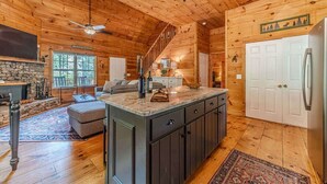 L’intérieur d’une cabane en rondins, avec une cheminée en pierre, un îlot de cuisine, un escalier et une grande fenêtre.