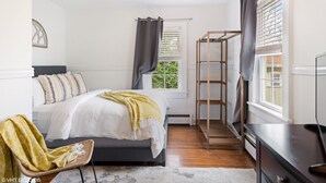 Un dormitorio con cama, una silla, un mueble para televisión y una ventana con cortinas.