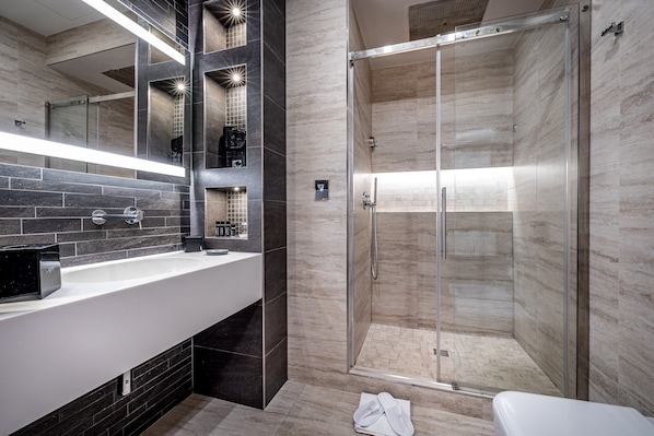 Un baño moderno con ducha acristalada, un lavabo blanco con dispensador de jabón negro e un espejo con iluminación integrada.
